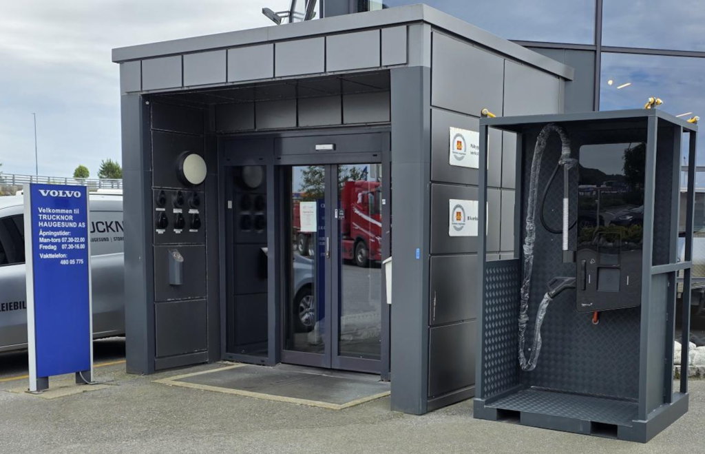Gray modular service kiosk with glass doors and hoses, next to a blue Volvo sign at a dealership or car wash area