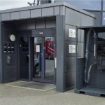 Exterior view of a gray service kiosk with a blue Volvo sign, glass doors, and a separate equipment cabinet with a coiled hose outside a glass building.