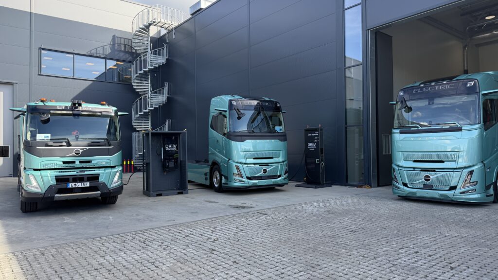Three teal Volvo electric trucks parked outside a modern industrial building with a spiral metal staircase visible on the wall.